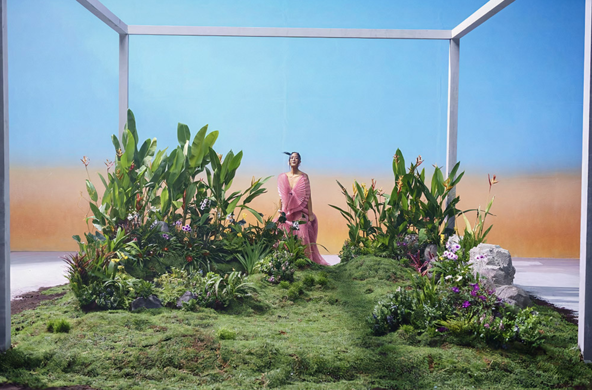 A Travipome scenography installation with plants, a performer, and a painted sky backdrop.